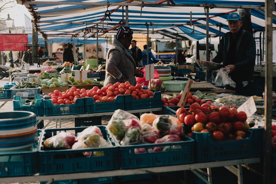 A bustling outdoor market scene under a large white canopy tent shows multiple vendors displaying various second-hand goods and household items on tables and shelves. In the foreground, a person weari