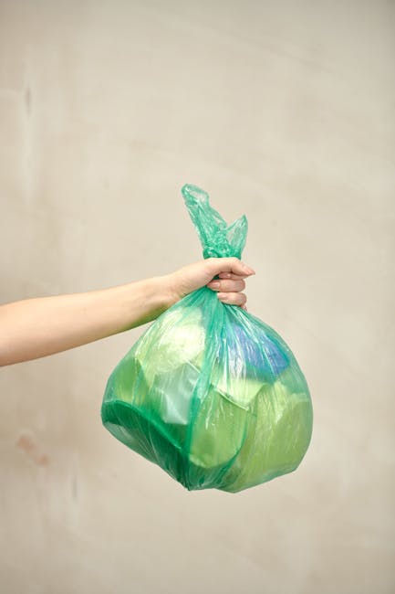 A human hand is holding a green and blue plastic bag, which appears to contain waste or rubbish. The bag is knotted at the top and is held by the fingers, with the arm extending from the left side of 