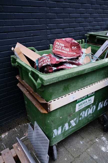 A human hand is holding a green and blue plastic bag, which appears to contain waste or rubbish. The bag is knotted at the top and is held by the fingers, with the arm extending from the left side of 