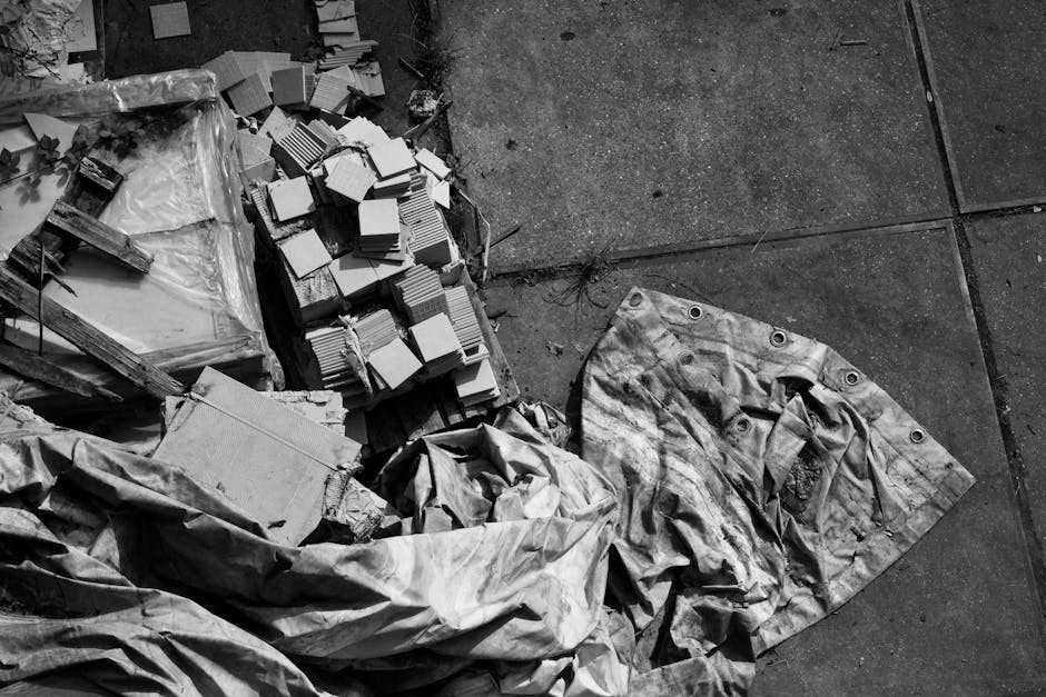 A man in a white shirt is seen lifting a large, heavily worn burlap sack filled with various waste items, including plastic bottles, containers, and tangled electrical wires. The sack appears aged and